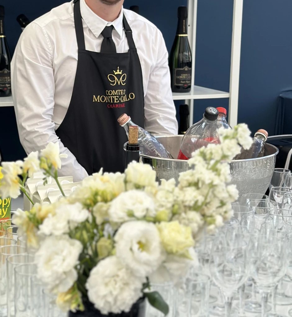 Tablier noir brodé or au logo d'une maison de champagne, sur un bar dressé de fleurs blanches